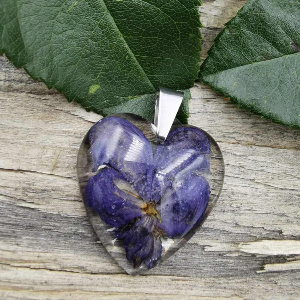 Wisiorek w kształcie serca zrobiony z przeźroczystej żywicy, w której utrwalono fioletowe kwiaty. Wisiorek wykończony jest minimalistyczną zawieszką w kolorze srebra.